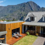 Villa devant les montagnes, avec son jardin et deux transats. On voit l'entrée de la maison avec son parking privé.